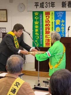 黄色いタスキを付けた市長が、黄緑色のジャケットを着て、タスキをかけた女性に、額入りの賞状を渡している写真