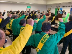 緑や、黄色、黄緑色のジャケットを着て、タスキを付けた交通安全の集いの参加者が、右手拳を挙げている写真