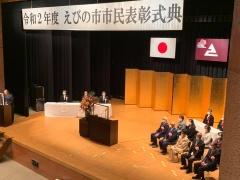 金屛風の置かれた舞台の右側に胸に花をつけた表彰者たちが座っている写真