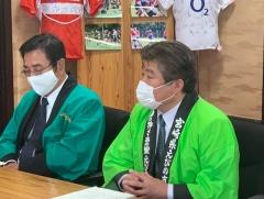 黄緑の法被を着てマスクをした市長と男性が座っている写真