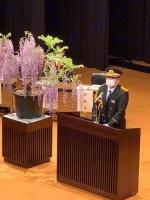 演台前に立つ正装した男性と、男性の左側に紫色の奇麗な花が置かれた写真