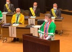 緑の法被を着た市長が演台前に立ち、その後方の議席に着席する関係者の写真