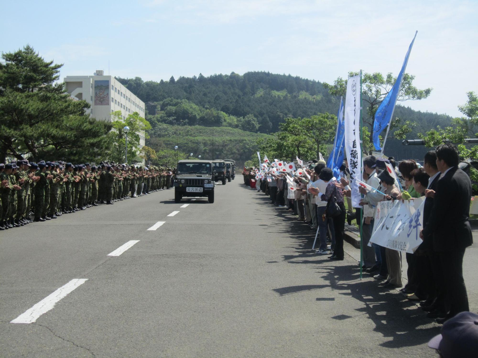 車両が通る道路わきに並ぶ迷彩服の部隊と、その向かいに旗や横断幕を持って並ぶ老若男女の写真