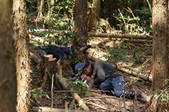 林の中で横たわっている帽子を被った男性の横に黒い犬の写真