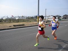 道路を赤いユニフォームと青いユニフォームを着た二名の駅伝選手が走っている写真