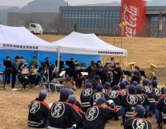 白いテントの下で、吹奏楽部の生徒が楽器を演奏しており、法被を着た消防団員が演奏を聞いている様子の写真