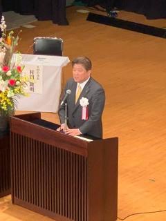 胸章リボンをつけた市長が、舞台上の演台に立って話をしている写真