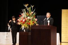 大きな花が飾られている舞台の講演台で市長が話をしている写真