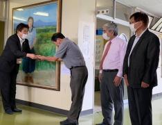 女性が描かれた絵画作品の前で市長が寄贈者の男性から目録を受け取っている写真