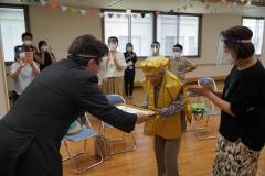 フェイスガードを付けた市長が金色のちゃんちゃんこを着た長寿者の女性に賞状を渡している写真