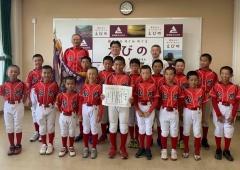 赤いユニフォーム姿の飯野亀城野球スポーツ少年団の選手達が市長と並んでおり、前列中央の男の子が賞状を持っている写真