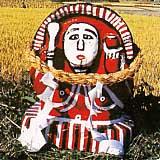 金色の稲穂を背に赤と白で塗られた田の神さぁの石像が田んぼに置かれている写真