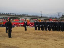 奥に駐車している消防車の右側に、整列した制服を着た消防団の人達のの写真