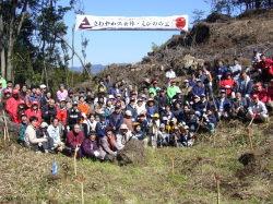 山の開けた場所で植樹祭の参加者が集まり記念撮影をしている写真