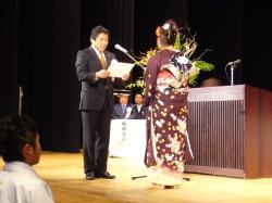 舞台上で振袖を着た女性の前で、市長が賞状を読み上げている写真
