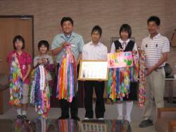市内保育園、小中学校から送られた千羽鶴と、額に入った賞状を持つ生徒4名と、市長、男性の記念写真