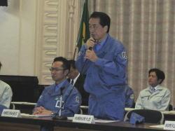 青い作業着を着た菅首相が両手でマイクを持って話をしている写真